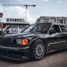 Czarny Mercedes 190 z agresywnym body kitem Evo, lśniącym na zlotowym placu.