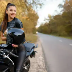 Uśmiechnięta dziewczyna z kaskiem w ręku opiera się o czarny motor. Idealny motor dla niskiej dziewczyny, gotowy na jesienną przejażdżkę.