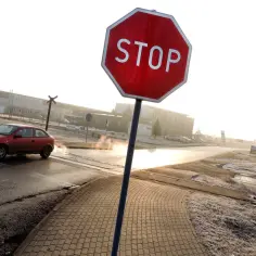 Czerwony samochód zbliża się do skrzyżowania, gdzie znak stop wskazuje, gdzie się zatrzymać. W tle budynki i kolej.